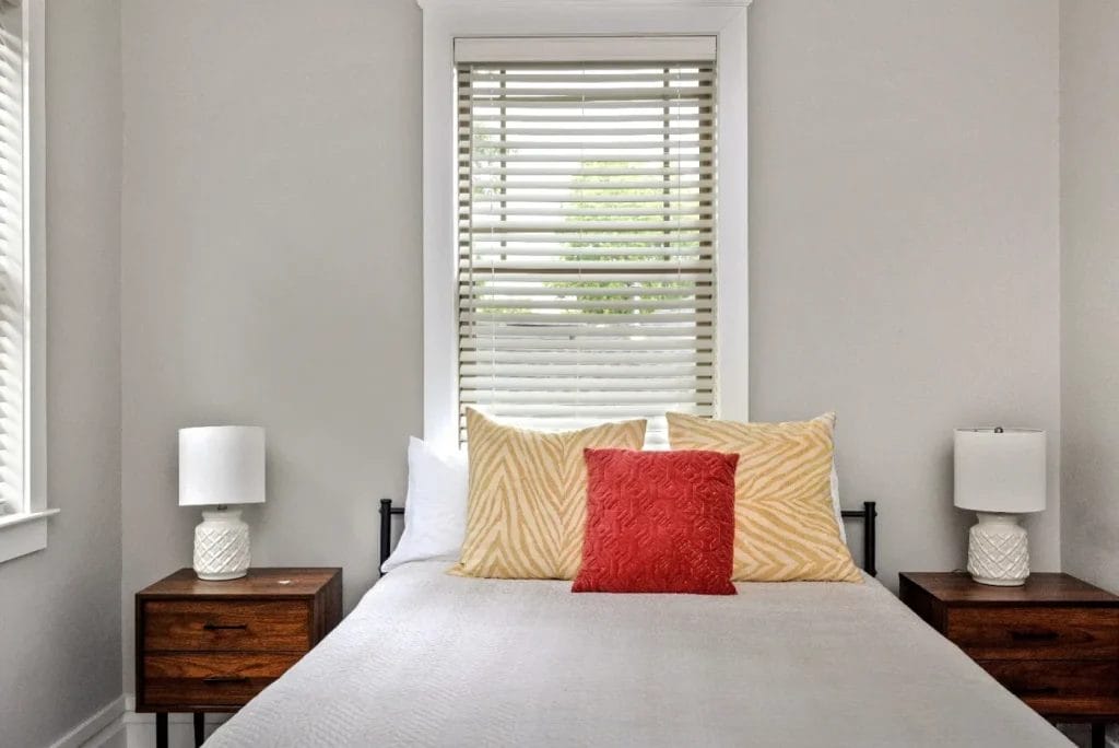A neatly made bed with two yellow patterned pillows, one red pillow, and a white bedspread sits between two wooden nightstands with white lamps. A window with blinds is centered above the bed.