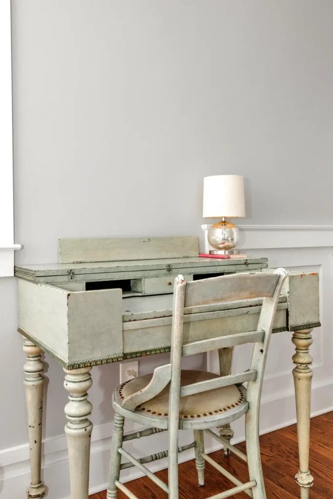 A vintage pale green wooden desk and matching chair are set against a light gray wall. A small table lamp with a round glass base and white shade sits on the back corner of the desk.
