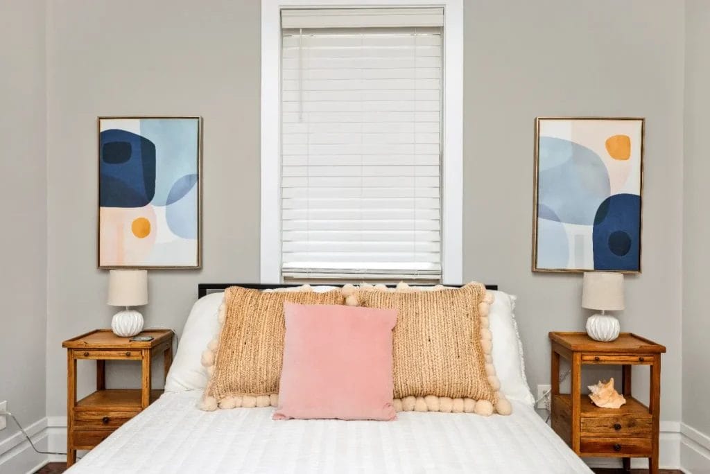 A neatly made bed with a pink cushion and tan textured pillow is placed between two wooden nightstands, each with a small lamp. Abstract art hangs on the light gray walls, and a window with closed blinds is above the bed.