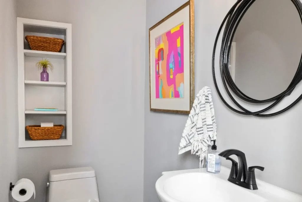 A modern bathroom with a white sink, black faucet, round black mirror, wall art with pink and yellow accents, a towel, shelving with wicker baskets, a small plant, and a toilet with toilet paper.