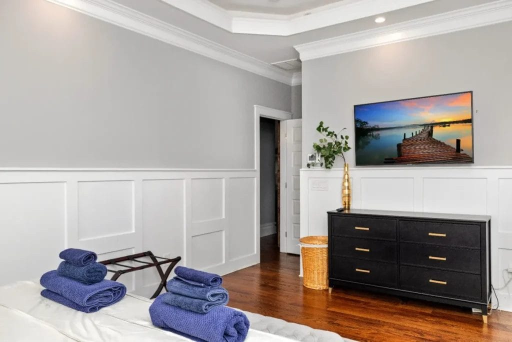 A modern bedroom features a neatly made bed with stacked blue towels, a black dresser with a plant and TV displaying a sunset over a dock, white wainscoting, hardwood floors, and a woven laundry basket.