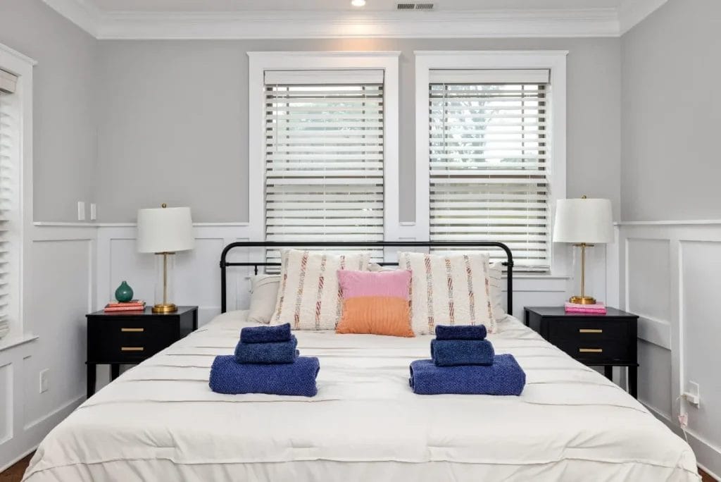 A neatly made bed with white bedding, colorful throw pillows, and two stacks of folded navy towels. Black nightstands with lamps and books flank the bed, and two windows with blinds are behind the headboard.