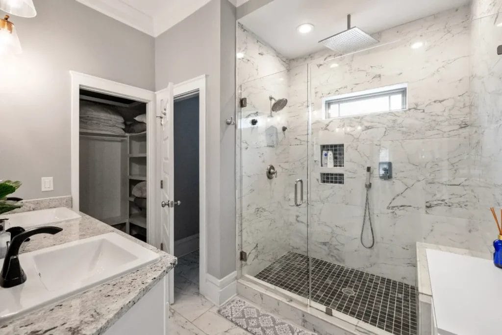 Modern bathroom with a glass-enclosed shower featuring marble walls and a rainfall showerhead, a vanity with a granite countertop and black faucet, and an open door revealing a walk-in closet with shelves.