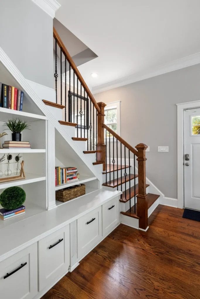 A staircase with wooden steps and black railings beside built-in white shelves and cabinets, decorated with books, plants, and baskets, on a hardwood floor in a bright entryway.