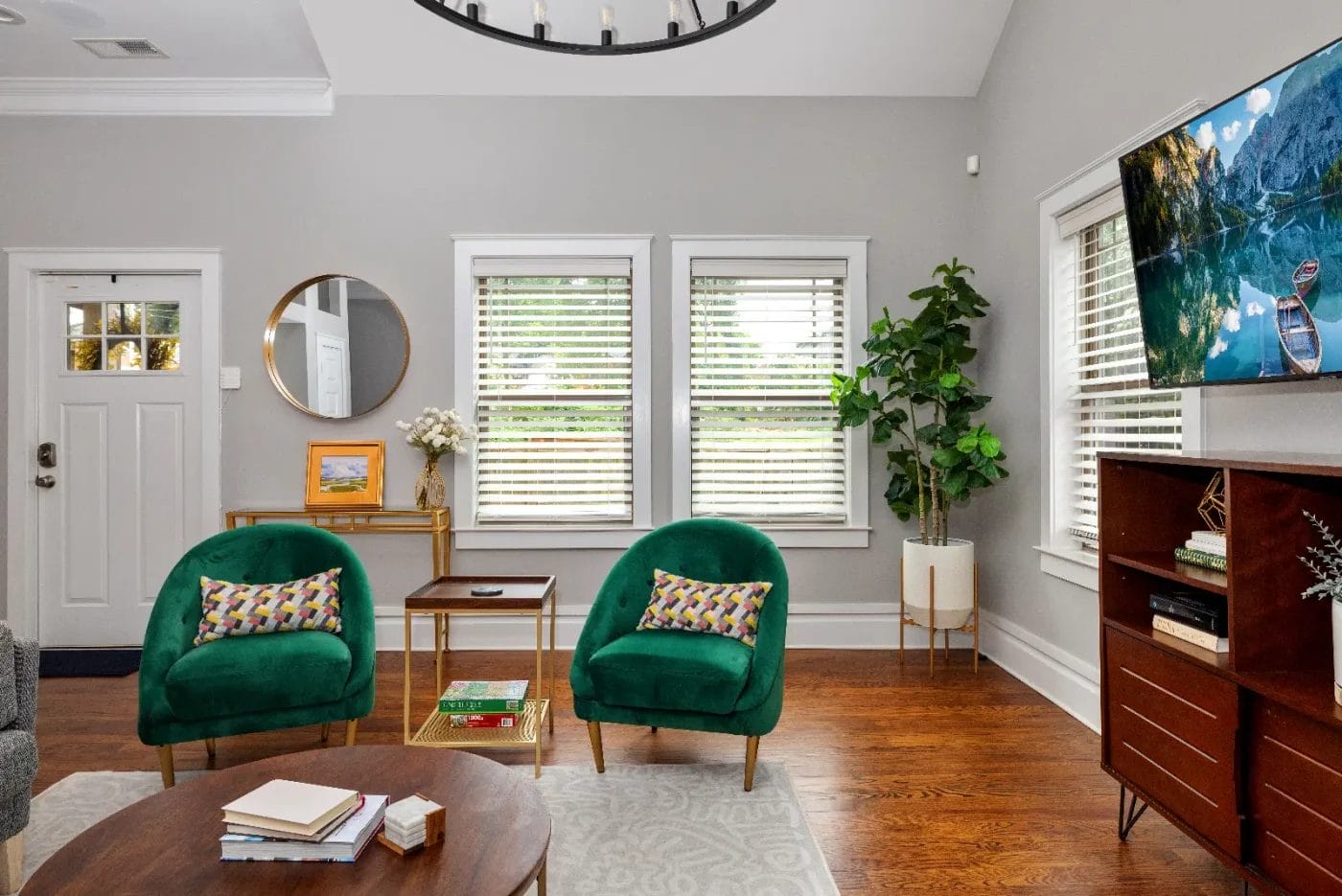 A modern living room with two green chairs, a round wooden coffee table, a TV on a wooden console, a large plant, and bright windows with blinds. Decor includes colorful pillows, books, and wall art.
