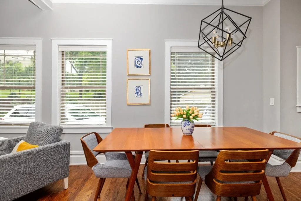A modern dining room with a wooden table, six chairs, a vase of yellow tulips, a gray armchair, three windows with blinds, framed art on the wall, and a geometric black chandelier.