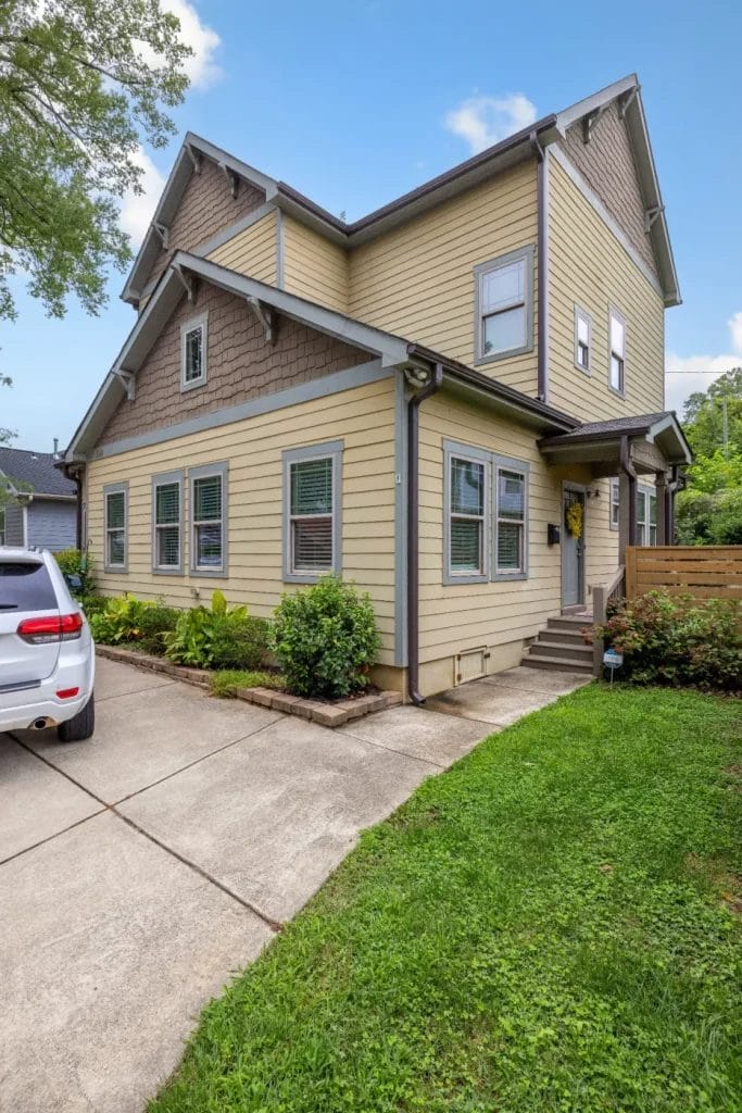 A two-story yellow house with gray trim and a gabled roof, featuring multiple windows, a small front porch, driveway, and a white SUV parked beside it. Lush green grass and shrubs line the walkway.