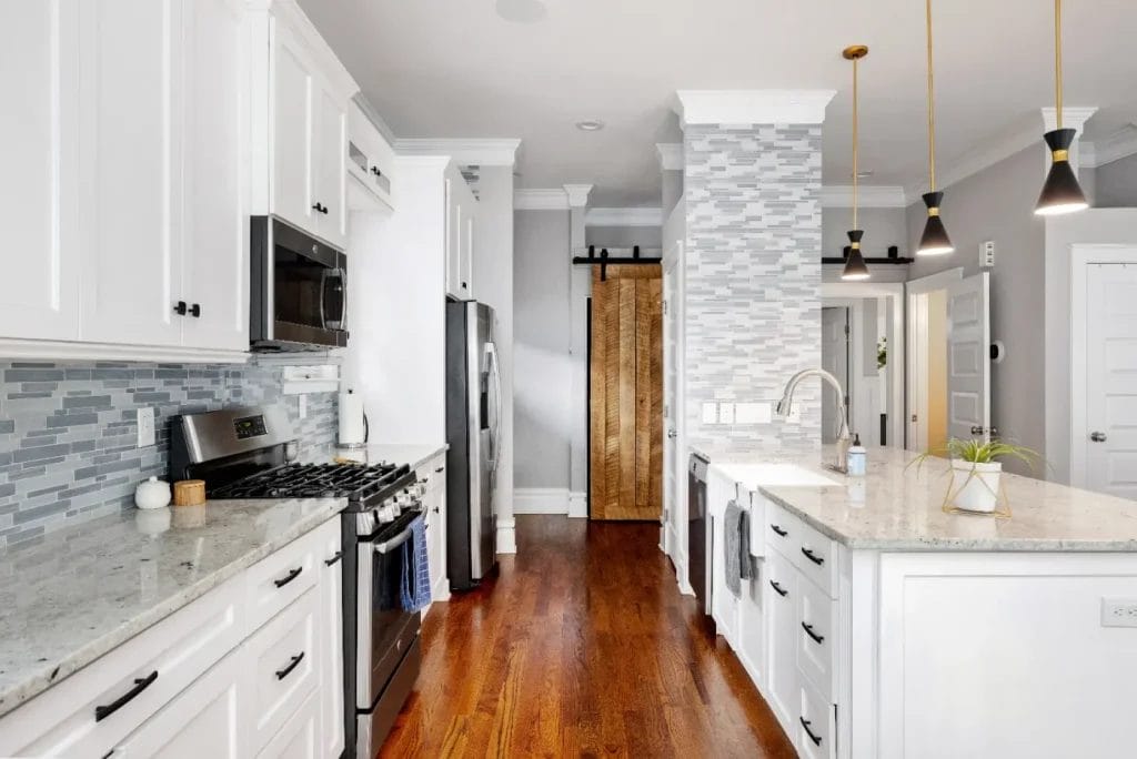 Modern kitchen with white cabinets, stainless steel appliances, a gray tile backsplash, marble countertops, wooden floors, pendant lights, and a sliding wooden barn door in the background.