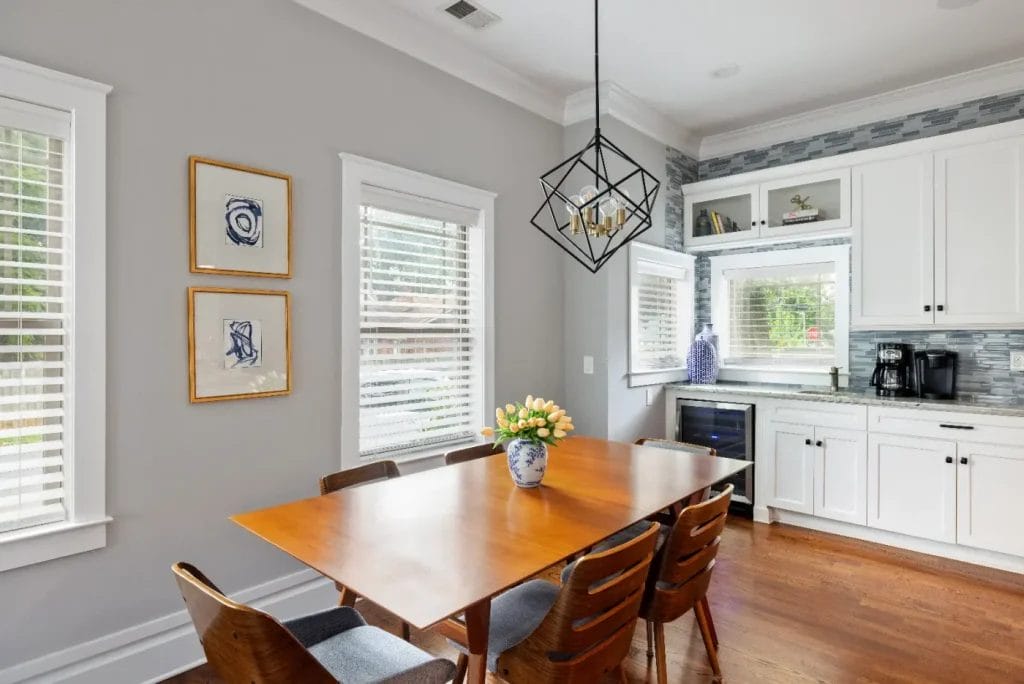 A modern dining area with a wooden table, four chairs, a vase of yellow flowers, white cabinets, coffee maker, wine fridge, and large windows with white blinds letting in natural light.