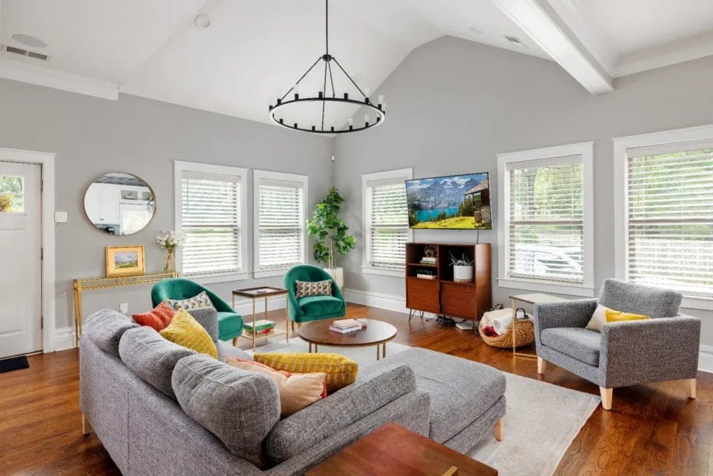 A bright, modern living room with large windows, gray couch, green armchairs, yellow accents, a round coffee table, wooden floor, TV, and indoor plants. White walls and exposed beams create an airy feel.