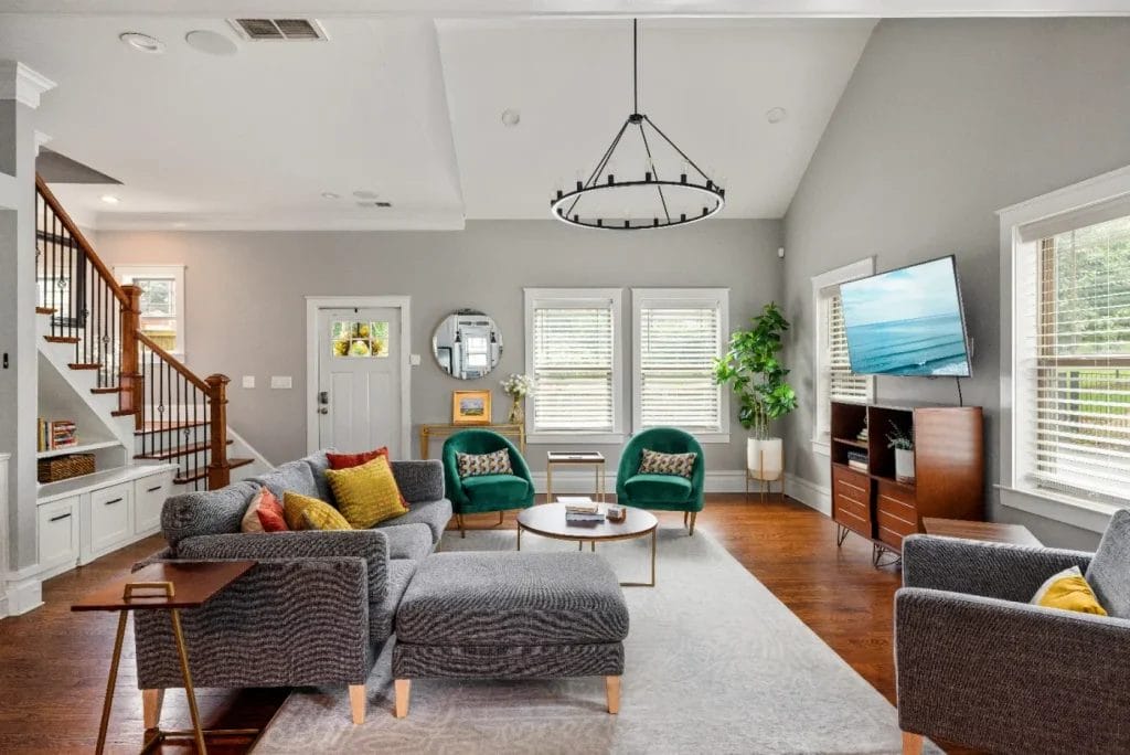 Bright, modern living room with gray sofas, green chairs, a round coffee table, TV on a wooden stand, large windows with blinds, indoor plant, and wooden floors. Stairs are visible to the left.