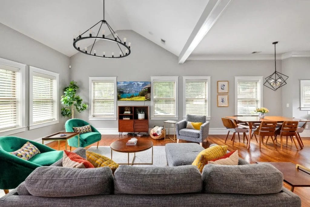 Bright, modern living and dining room with large windows, gray sofa, green chairs, round coffee table, TV on console, indoor plant, and wooden dining table with chairs on a hardwood floor.