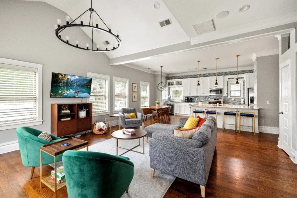 Modern open-concept living room and kitchen with gray sofa, green chairs, TV, hardwood floors, and a large chandelier. The kitchen features bar stools, pendant lights, and white cabinetry. Bright natural light fills the space.