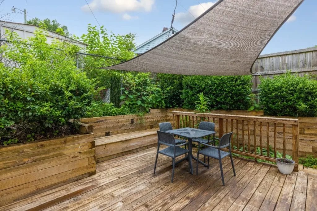 A wooden patio with a small table and four chairs, surrounded by lush greenery and wooden planters. A fabric shade sail is stretched overhead, providing partial shade to the seating area.