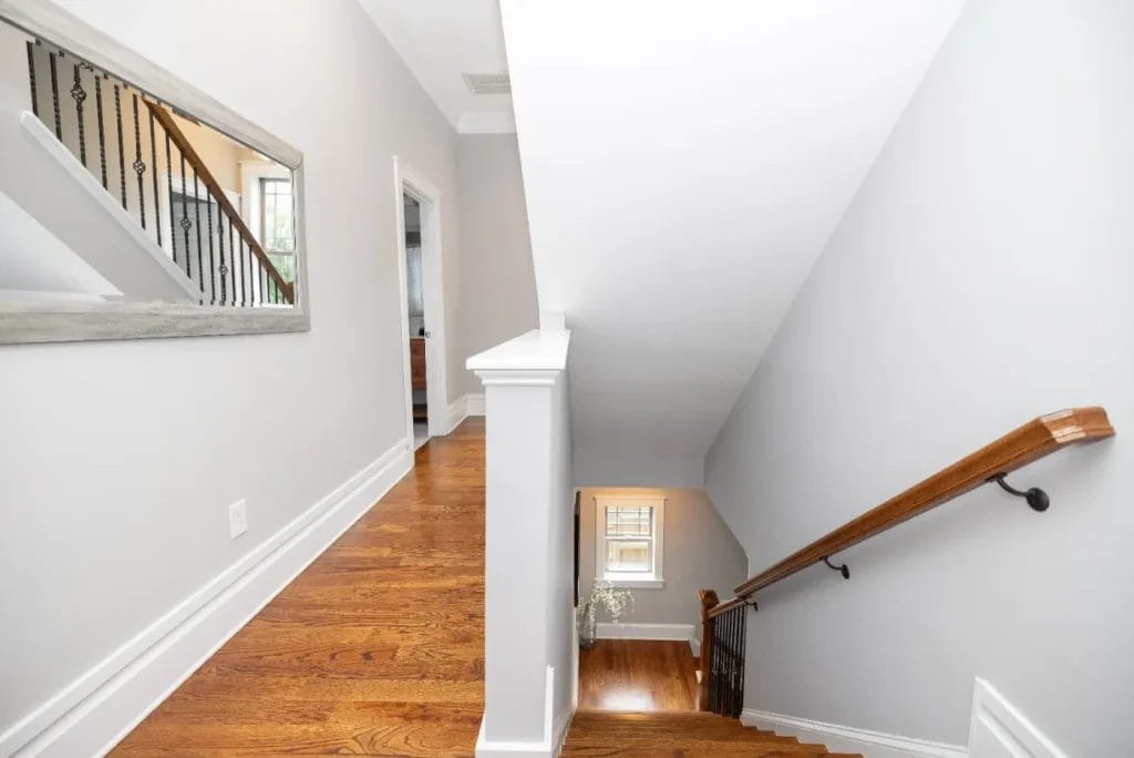 Bright hallway with light gray walls, hardwood floor, a large wall mirror on the left, and a staircase with a wooden handrail and black balusters descending to a lower level.