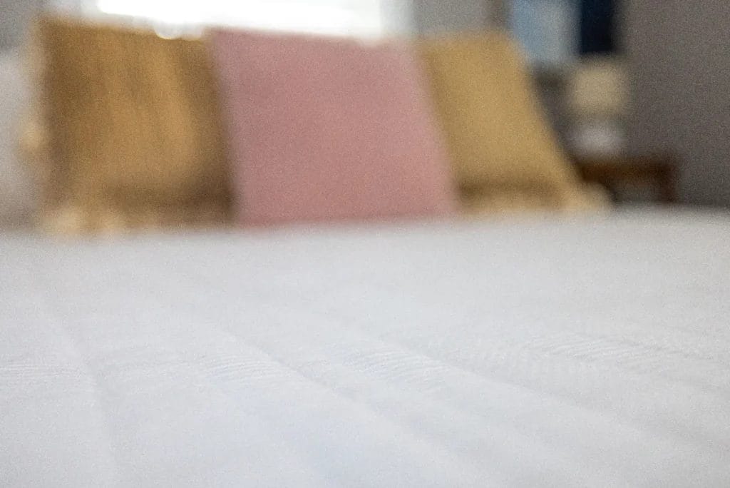 A soft focus photo of a neatly made bed with a white textured blanket and three out-of-focus pillows—two tan on the sides and one pink in the middle—against a blurred background.
