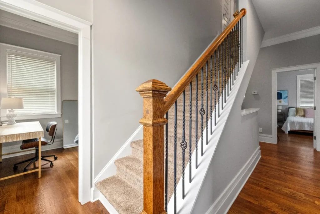 A carpeted staircase with a wooden handrail and black iron balusters is next to a hallway with hardwood floors. A small office with a desk and lamp is on the left, and a bedroom is visible in the background on the right.