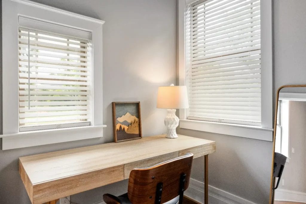 A minimalist home office features a wooden desk, a dark wood chair, a white table lamp, a framed mountain artwork, a tall mirror, and two windows with white blinds letting in natural light.