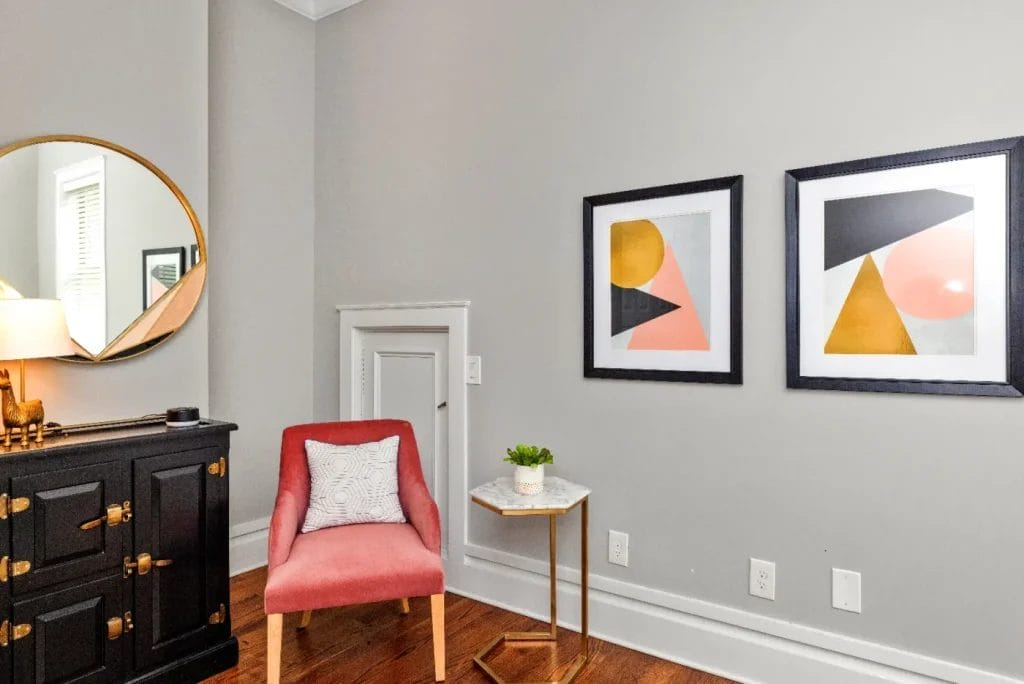 A modern room corner with a pink chair, white pillow, small marble side table with a plant, black dresser, round mirror, and two framed abstract art pieces on a light gray wall.