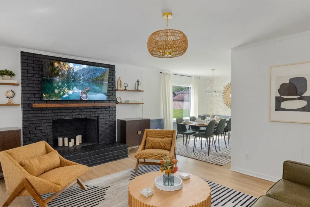 A modern living room with mustard yellow chairs, a round wooden coffee table, a black brick fireplace with candles, a large wall-mounted TV, and a dining area with dark chairs and a large window in the background.