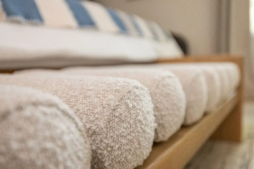 Close-up of neatly rolled white towels stacked on a wooden shelf, with a blurred background featuring a bed with blue and white striped pillows.