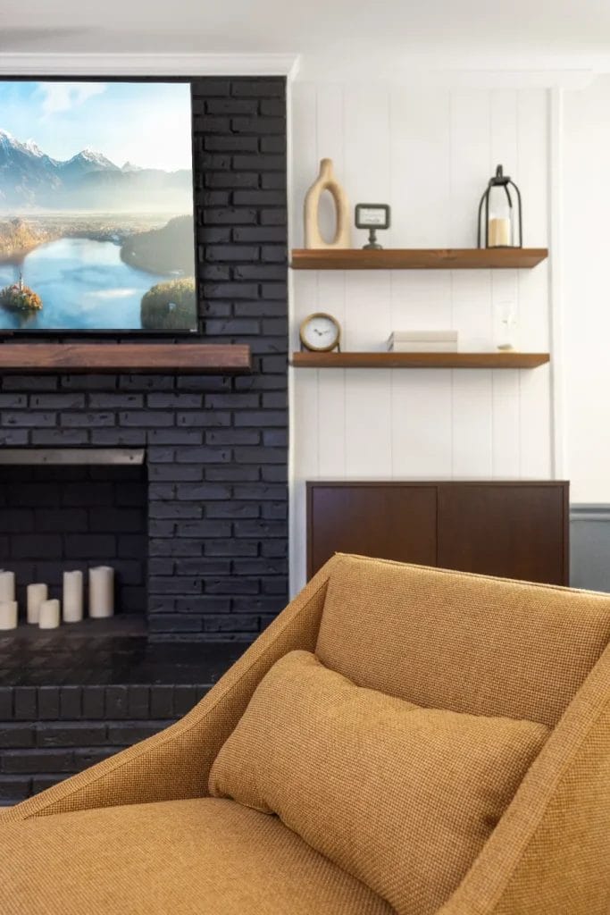 A cozy living room corner with a mustard-colored armchair, a black brick fireplace with candles, a TV above, and wooden shelves holding decor, a clock, and glassware on a white wall.