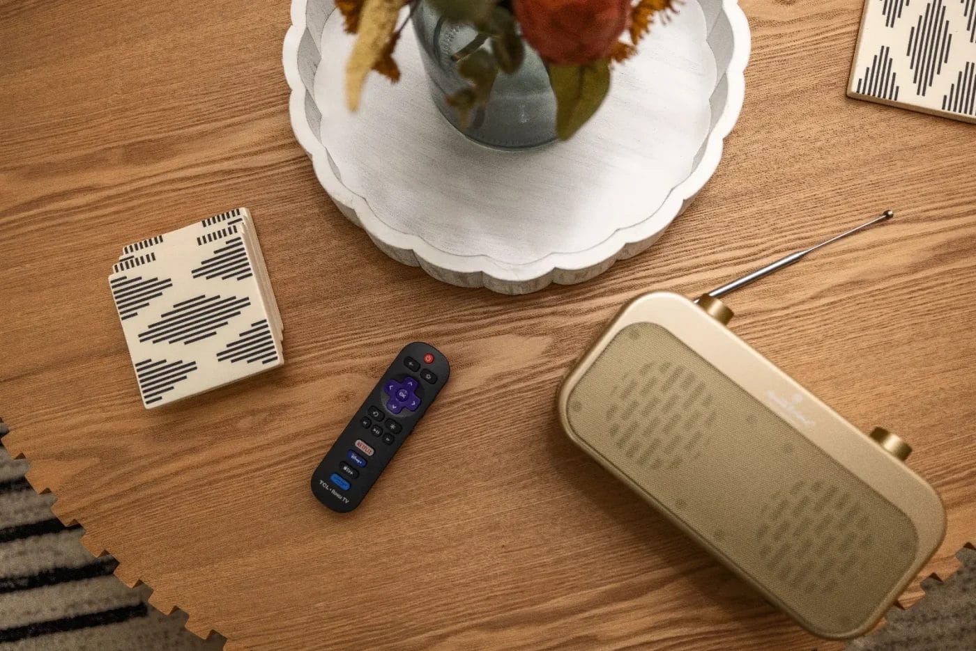 A wooden table with a patterned ceramic box, a black TV remote, a retro beige radio with an antenna, and a round tray holding a vase of flowers.