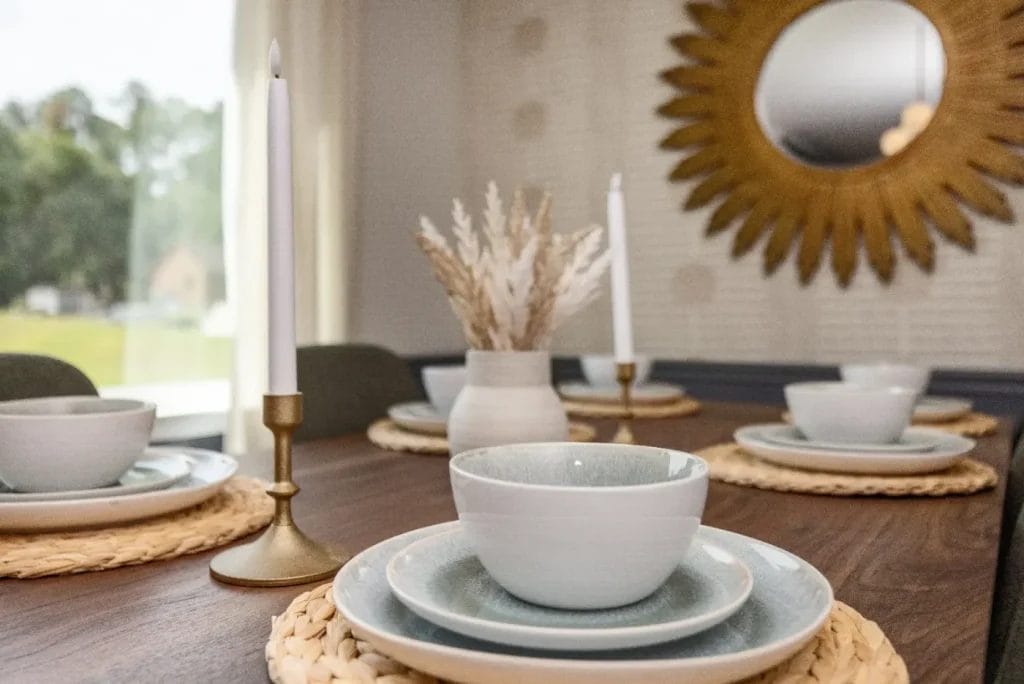 A wooden dining table set with white plates, bowls, and gold candlesticks, arranged on woven placemats. A vase with dried grasses is in the center, and a sunburst mirror hangs on the wall in the background.