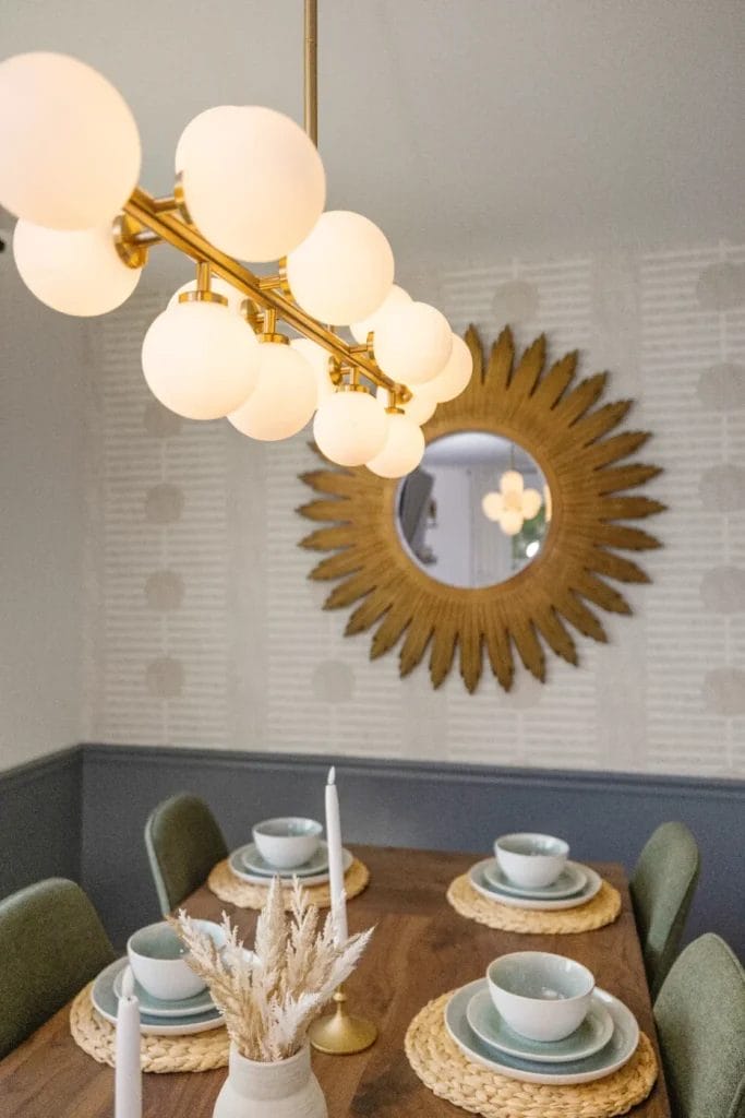 A modern dining room with a wooden table set for four, featuring woven placemats, pastel dishes, and white candles. A gold sunburst mirror and globe pendant lights hang above, reflecting the room’s cozy, stylish decor.
