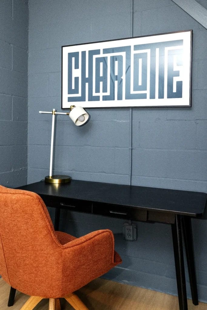 A modern workspace with an orange chair, a black desk, a gold and white desk lamp, and a framed geometric artwork spelling "CHARLOTTE" on a gray-blue wall.