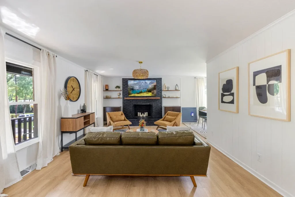 A modern living room with a green sofa, two tan chairs, a black fireplace, abstract art on white walls, light wood floors, and large windows with white curtains. A clock and TV are above the mantel.