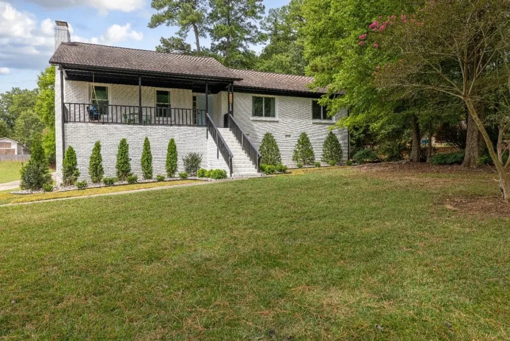 A white brick single-story house with a covered front porch, black railing, and a staircase leading to the entrance. The yard is grassy with neatly trimmed bushes and tall trees surrounding the property.