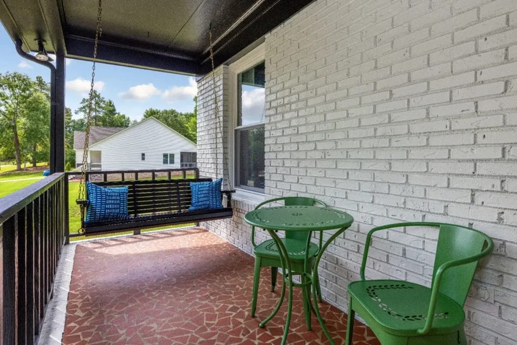 A cozy porch with a black wood swing, two blue pillows, and a small green metal table with two matching chairs. The floor is tiled, and the white brick wall is next to a window. Trees and a house are visible in the background.