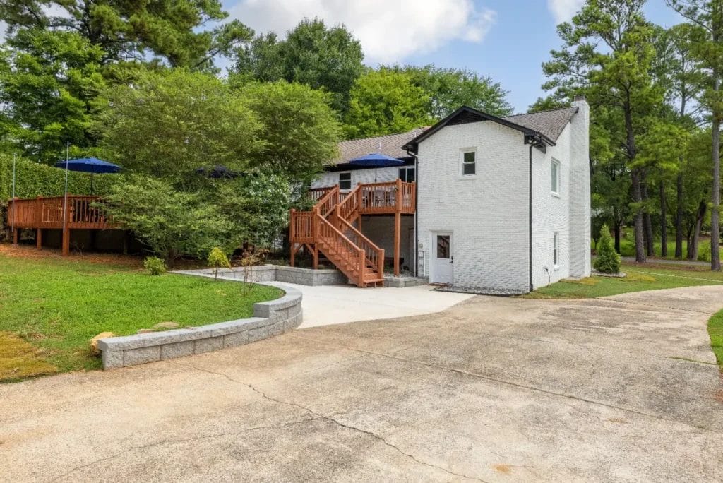 A white two-story house with a large wooden deck, surrounded by trees and greenery. A curved concrete driveway leads up to the house, and blue patio umbrellas are visible on the deck.