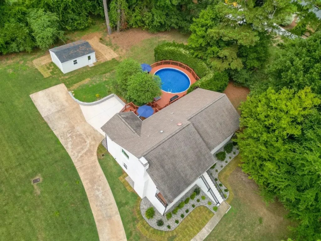 Aerial view of a house with a gray roof, a curved driveway, landscaped walkway, small detached shed, and a round above-ground pool on a wooden deck surrounded by trees and green lawn.