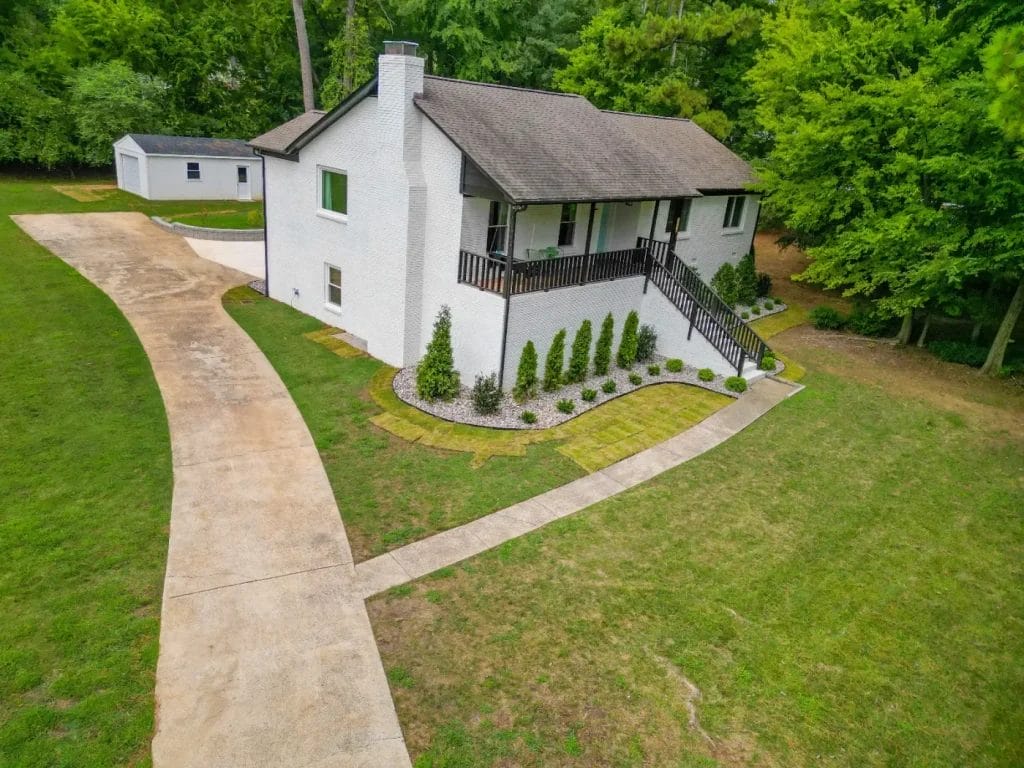 A white brick house with a covered front porch, dark railings, tidy landscaping, and a long curved driveway on a spacious grassy lot with trees surrounding the property.