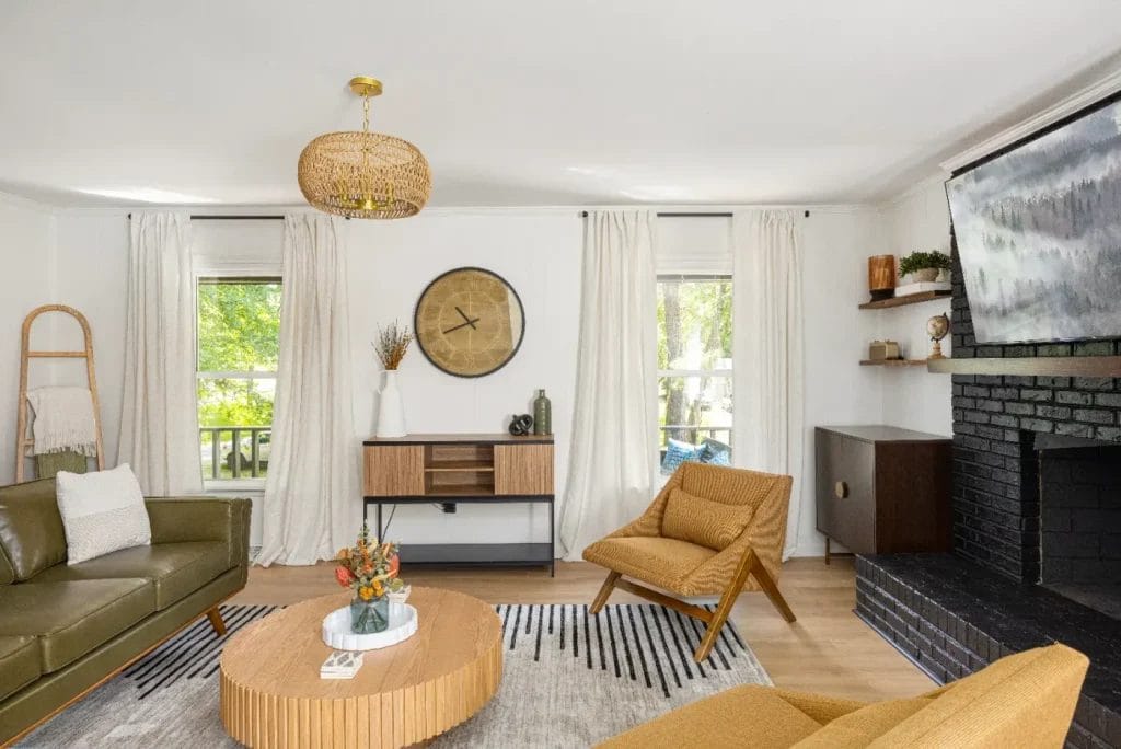 A modern living room with a green sofa, mustard armchair, round wooden coffee table, wall-mounted TV above a black brick fireplace, large clock, and two windows with white curtains letting in natural light.