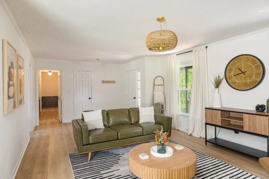 A modern living room with a green leather sofa, round wooden coffee table, black and white striped rug, wooden console, and wall art. Natural light enters through a window with white curtains.