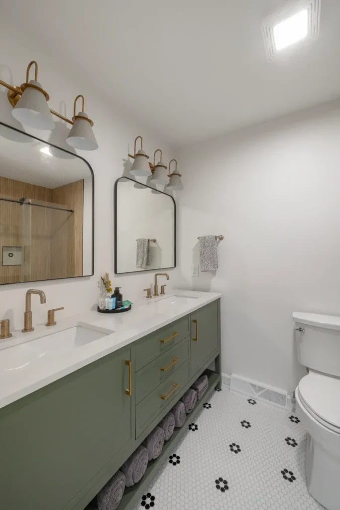 Modern bathroom with a double sink vanity in sage green, gold hardware, two mirrors, and three wall sconces. White walls, hexagon tile floor with black accents, and a toilet are visible. Towels stored on a shelf under the sinks.