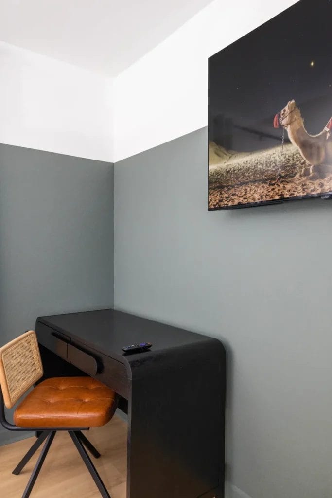 A modern minimalist workspace with a black desk, brown cushioned chair, and a remote control on the desk. A playful wall art featuring a camel in a desert scene hangs above on the green-gray wall.