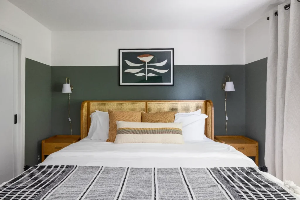 A neatly made bed with a wicker headboard, white and tan pillows, and a patterned blanket. Two bedside tables with wall-mounted lamps are on either side, and a framed abstract flower art hangs above the bed on a green and white wall.