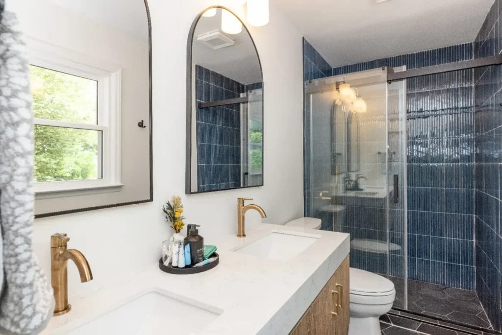 Modern bathroom with dual sinks, gold fixtures, a large mirror, a white countertop, blue tiled shower with glass doors, and a window letting in natural light. Towels and toiletries are neatly arranged on the counter.