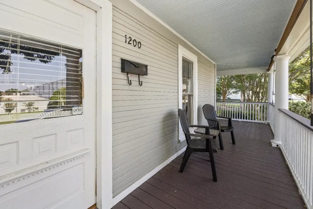 A front porch with two black chairs, a white door with blinds, house number 1200, beige siding, and a mailbox; the porch has dark wood flooring and white railings with greenery visible outside.