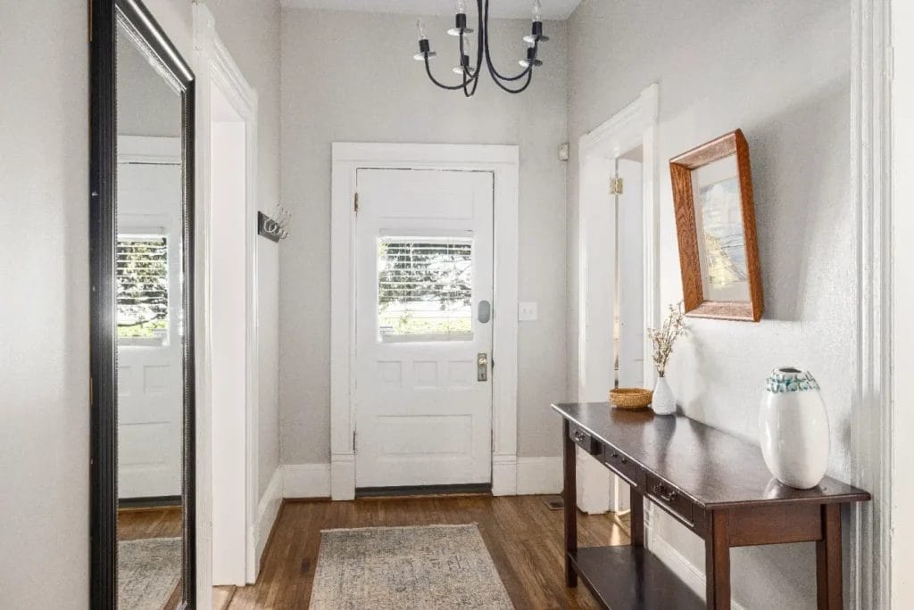 Bright hallway with a white front door, a large wall mirror on the left, a console table with decor on the right, wood floors, light gray walls, and a black chandelier overhead.