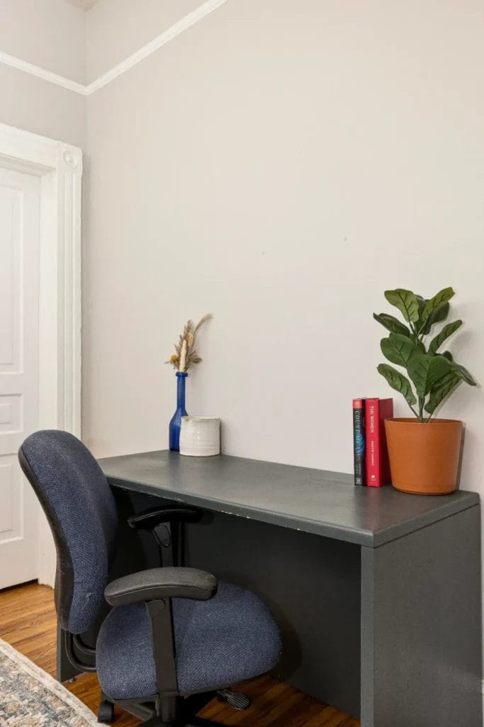 A simple home office setup with a gray desk, blue office chair, a potted plant, two books, a ceramic vase, and a blue glass vase with dried flowers against a light-colored wall.