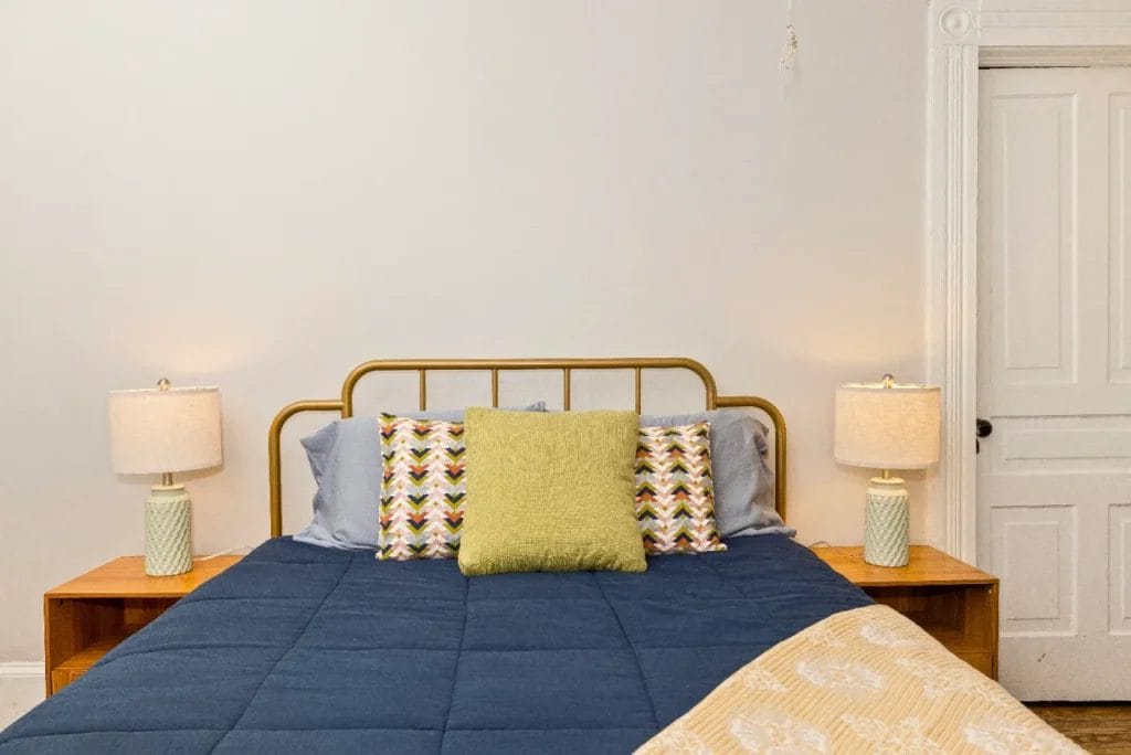 A neatly made bed with a blue comforter, decorative pillows, and two wooden nightstands. Each nightstand holds a lamp with a cream shade. The background features a white wall and a closed white door.