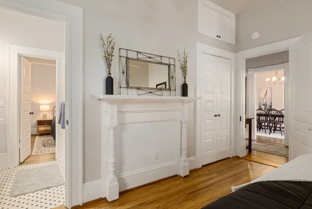 A bright bedroom with hardwood floors, white walls, a decorative white fireplace mantel with a mirror and vases, multiple doors, and a view into a dining room with a chandelier and black chairs.