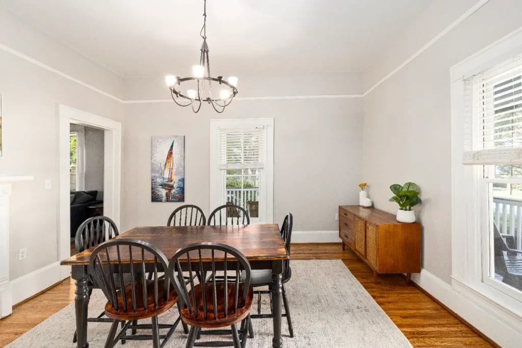Bright dining room with a dark wooden table, six chairs, a modern chandelier, a sideboard with plants and decor, a sailboat painting, hardwood floors, and large windows letting in natural light.