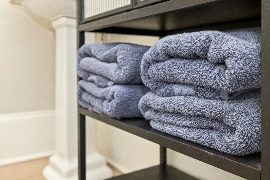 Four neatly folded blue towels stacked on two black metal shelves in a bathroom, with part of a white sink pedestal visible in the background.