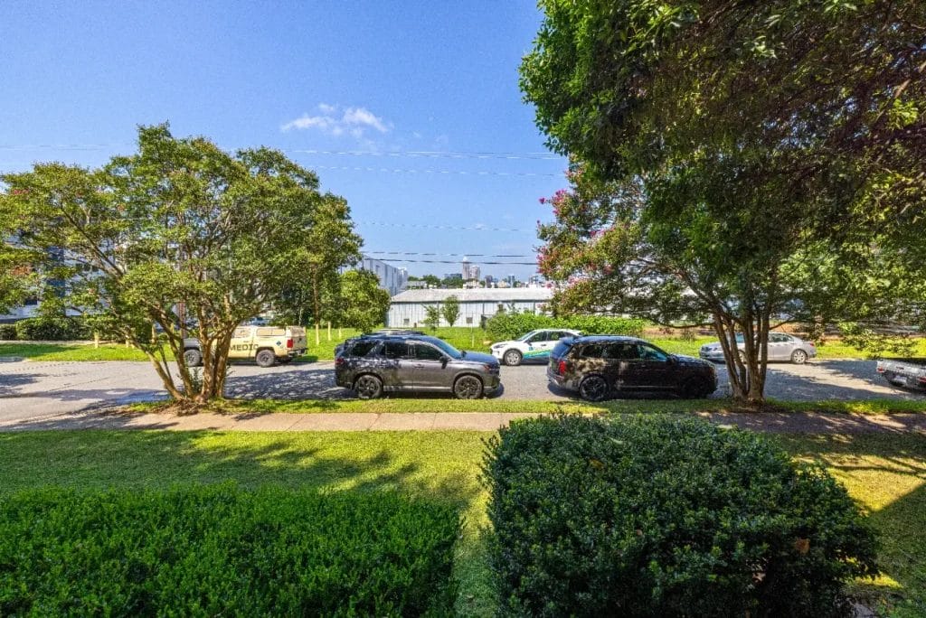 A sunny suburban street with several parked cars, green trees and bushes, and a white building in the background under a clear blue sky.